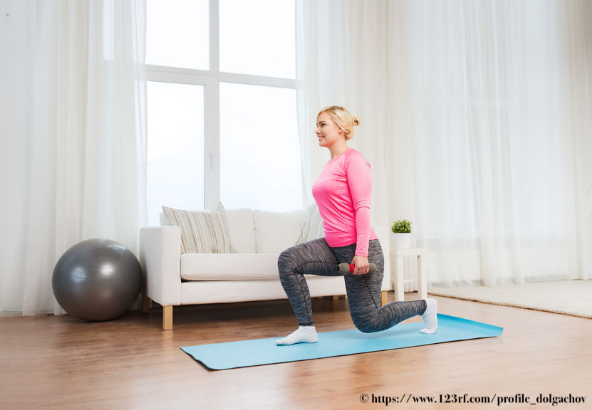 Blond, white woman in a pink long sleeve workout shirt and grey leggings performs a split squat with dumbbells. She is on a teal yoga mat that is on a hardwood floor. In the background it a large window with white walls, a white couch and a grey exercise ball.
